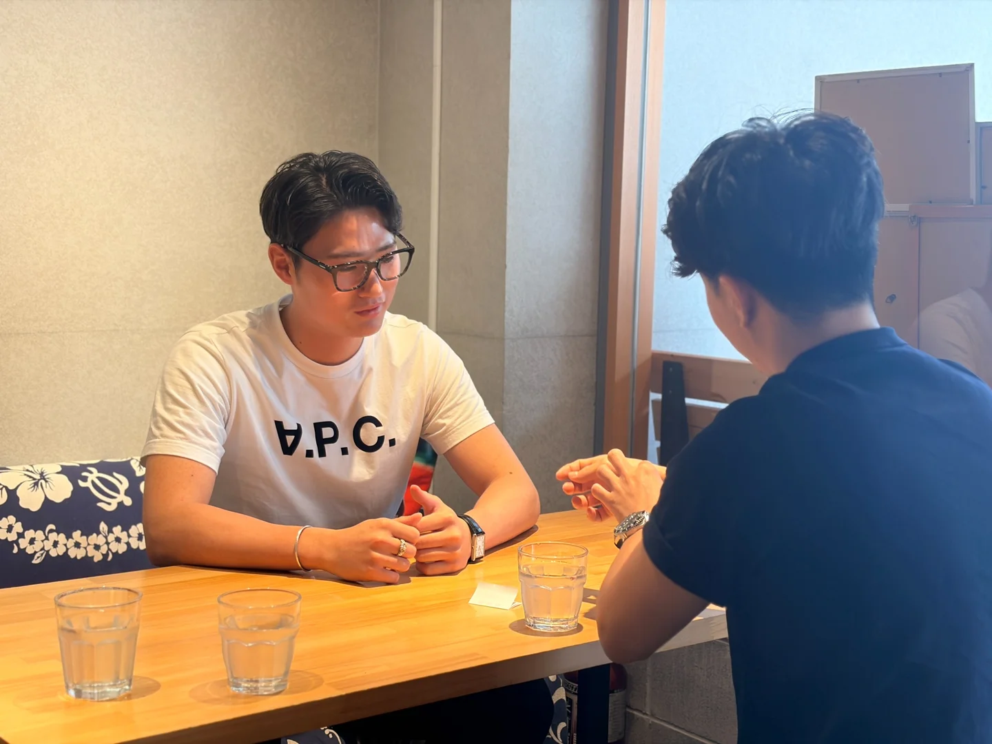 木のテーブルをはさんで、私服の学生2人が話している。眼鏡の学生が相手の話を聞いている。